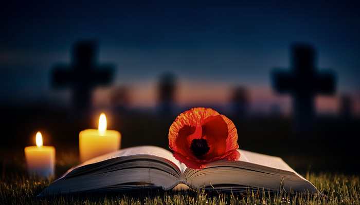 A single poppy lies on a book of remembrance in a military graveyard at night with candles illuminating the scene.