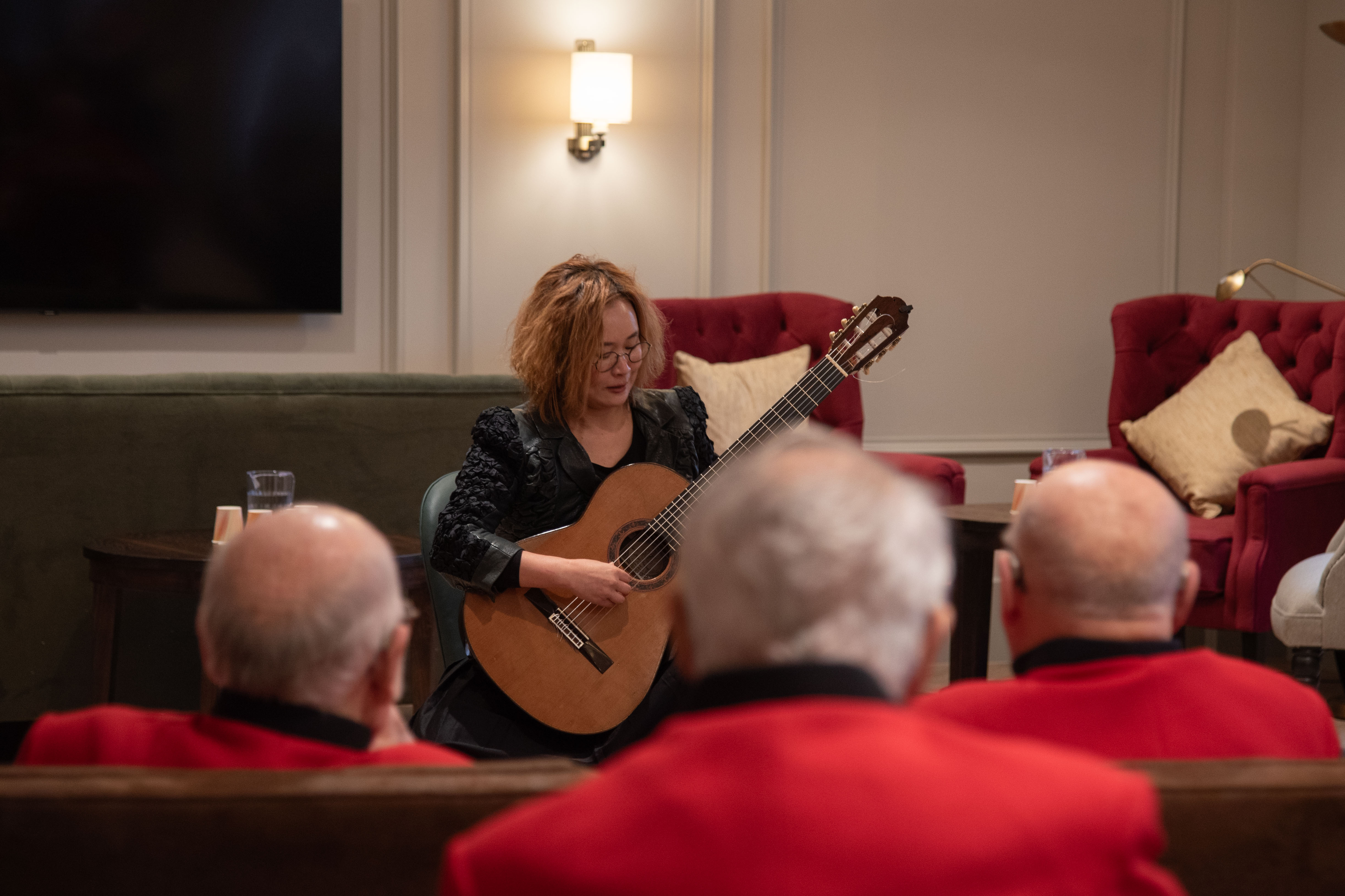 Musician Emily Yeyoung performs guitar watched by a group of Chelsea Pensioners in scarlet coats