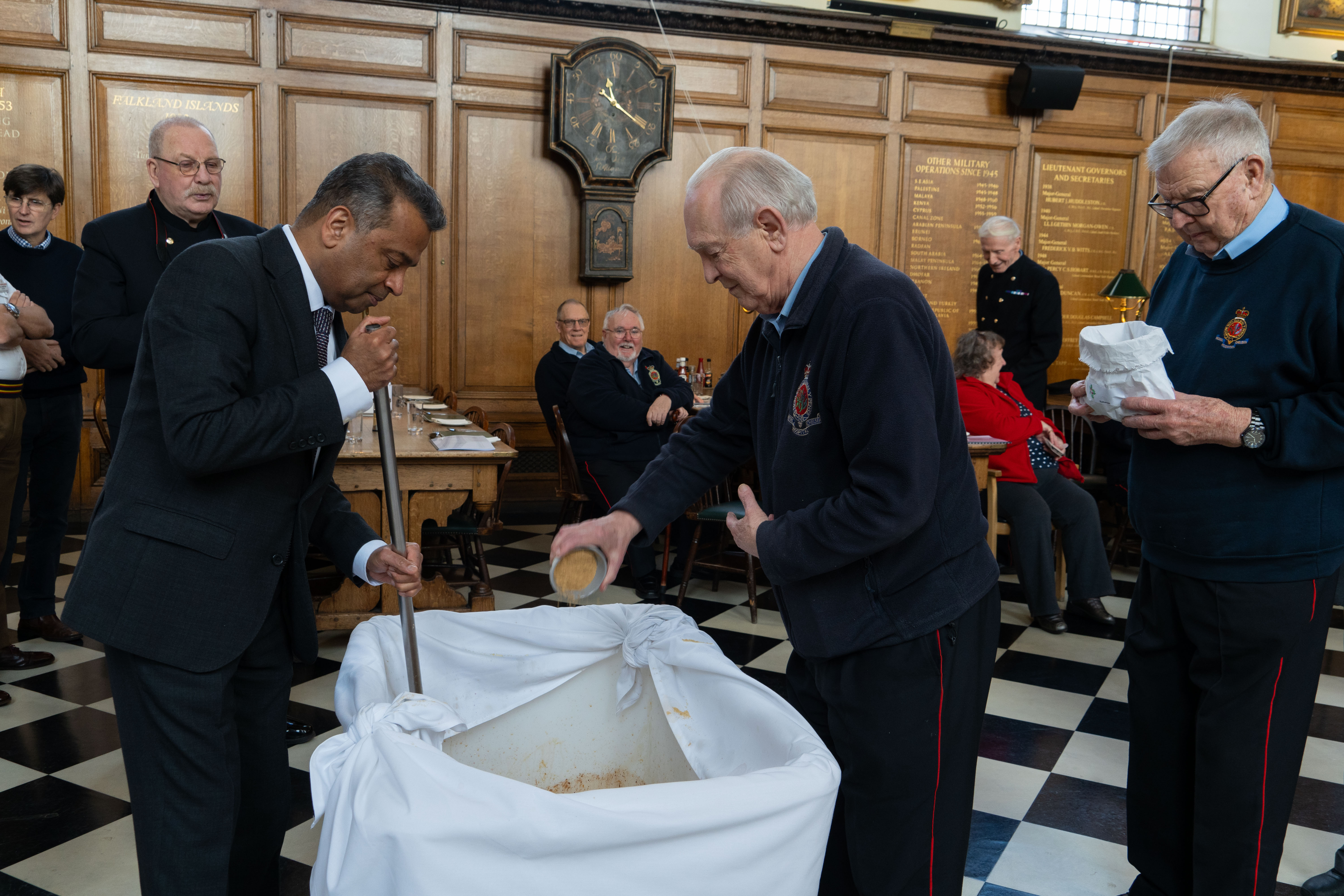 Chelsea Pensioner adding ingredients into large mixing bowl whilst someone stirs the mixture