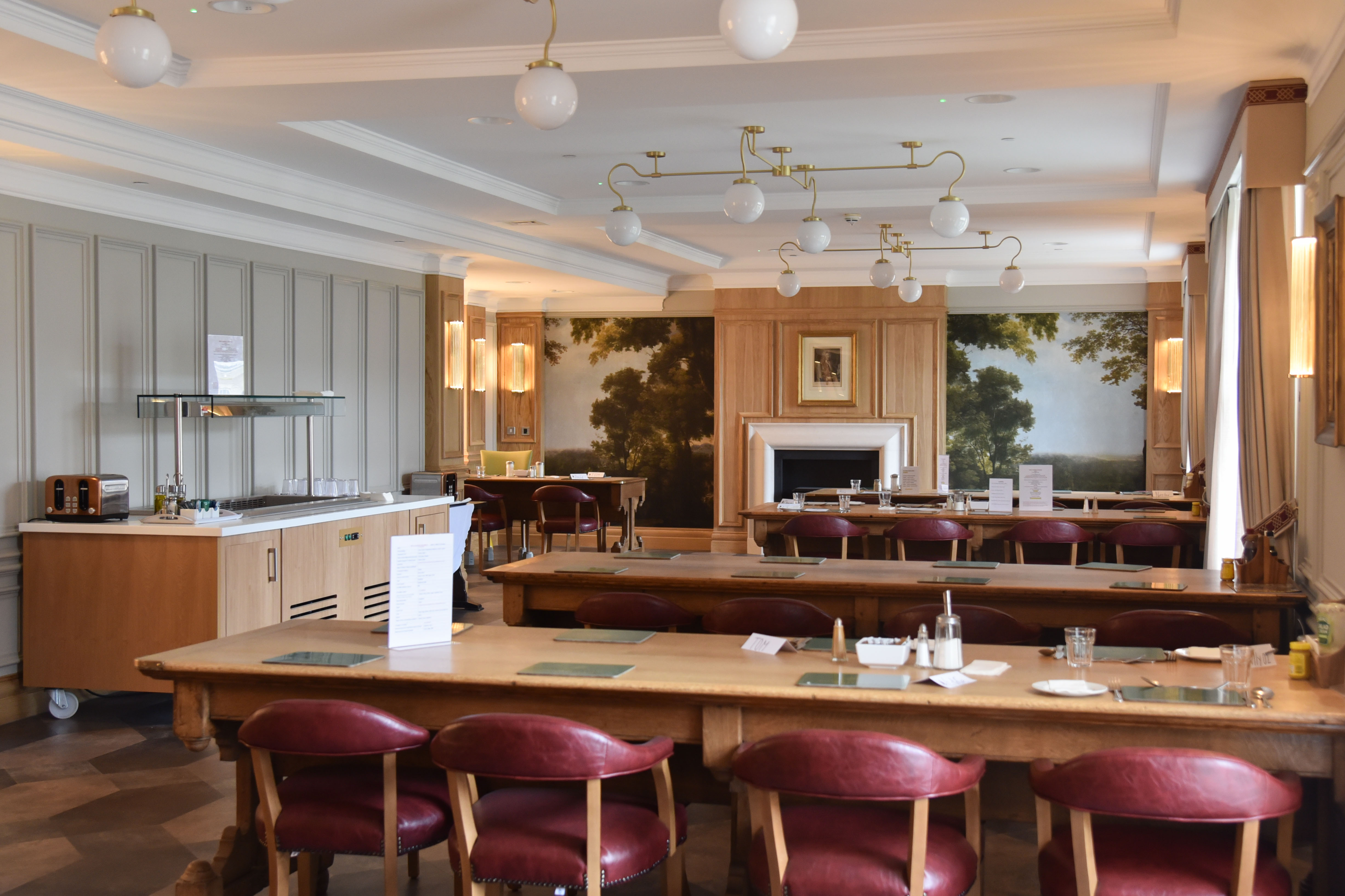 Dining space with long tables and chairs and tree painting and fireplace in the background