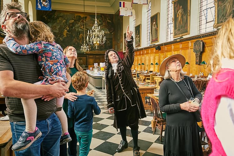 An actor dressed in period costume and representing Sir Christopher Wren stands inside the Great Hall at the Royal Hospital Chelsea with wood-panelled walls and chequered black and white floor tiles. A group of visitors of various ages listen in