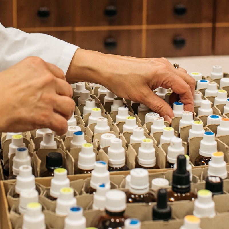 Hands selecting from a number of small vials with white lids. A series of small drawers in the background.