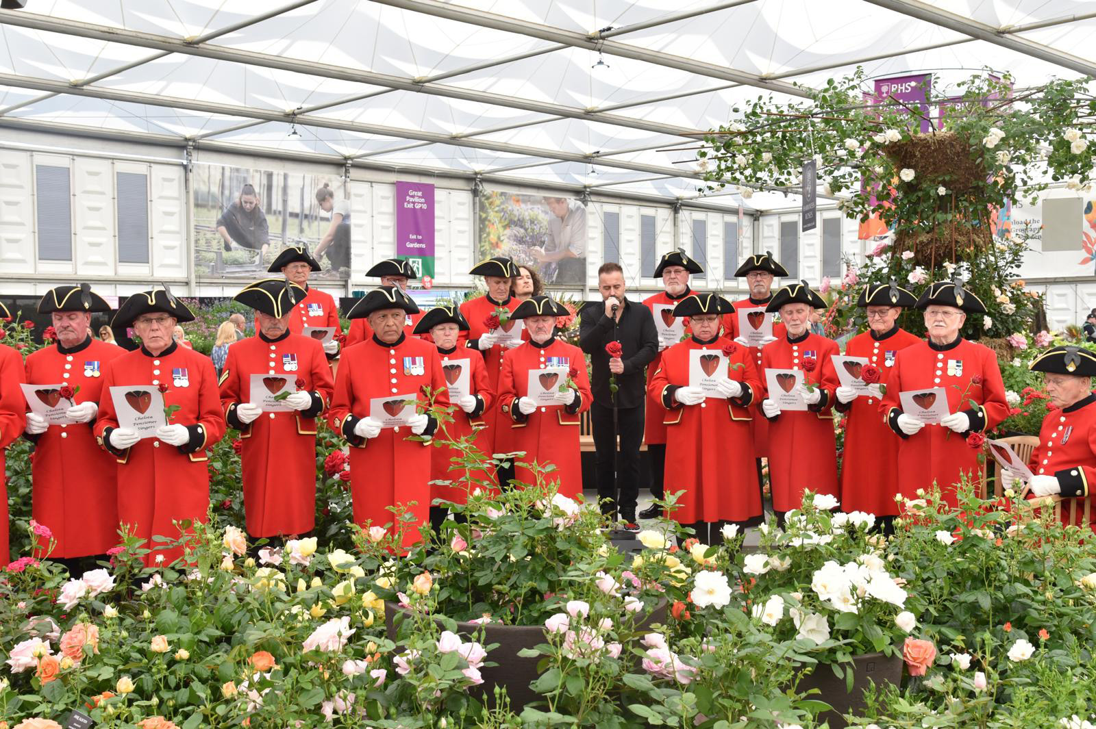 A group of Chelsea Pensioner Signgers stand with music books amongst flowers at the Chelsea Flower Show.