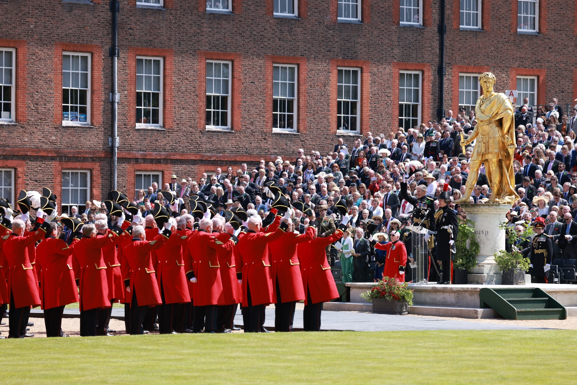 A group of Chelsea Pensioners lift their tricorne hats in salute to Princess Anne in front of a large crowd on a bright day during Founders Day 2024