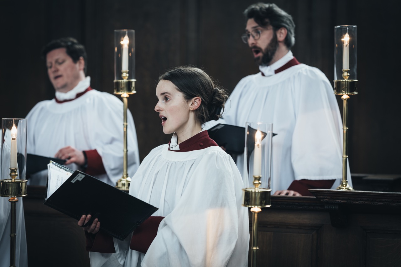 Two male and one female sing in traditional white church robes with maroon collars and sleeves indoors by candlelight