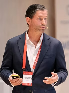 A suited guest speaker, Christopher Siedentopf, is shown talking, wearing a microphone and holding a remote control. A Red lanyard hangs around his neck.