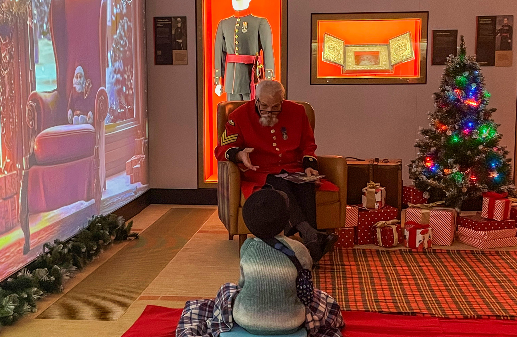 A Chelsea Pensioners sits in front of a young child in a Christmas themed grotto. A Christmas tree and lights twinkle in the corner and a digital backdrop extends the theme.