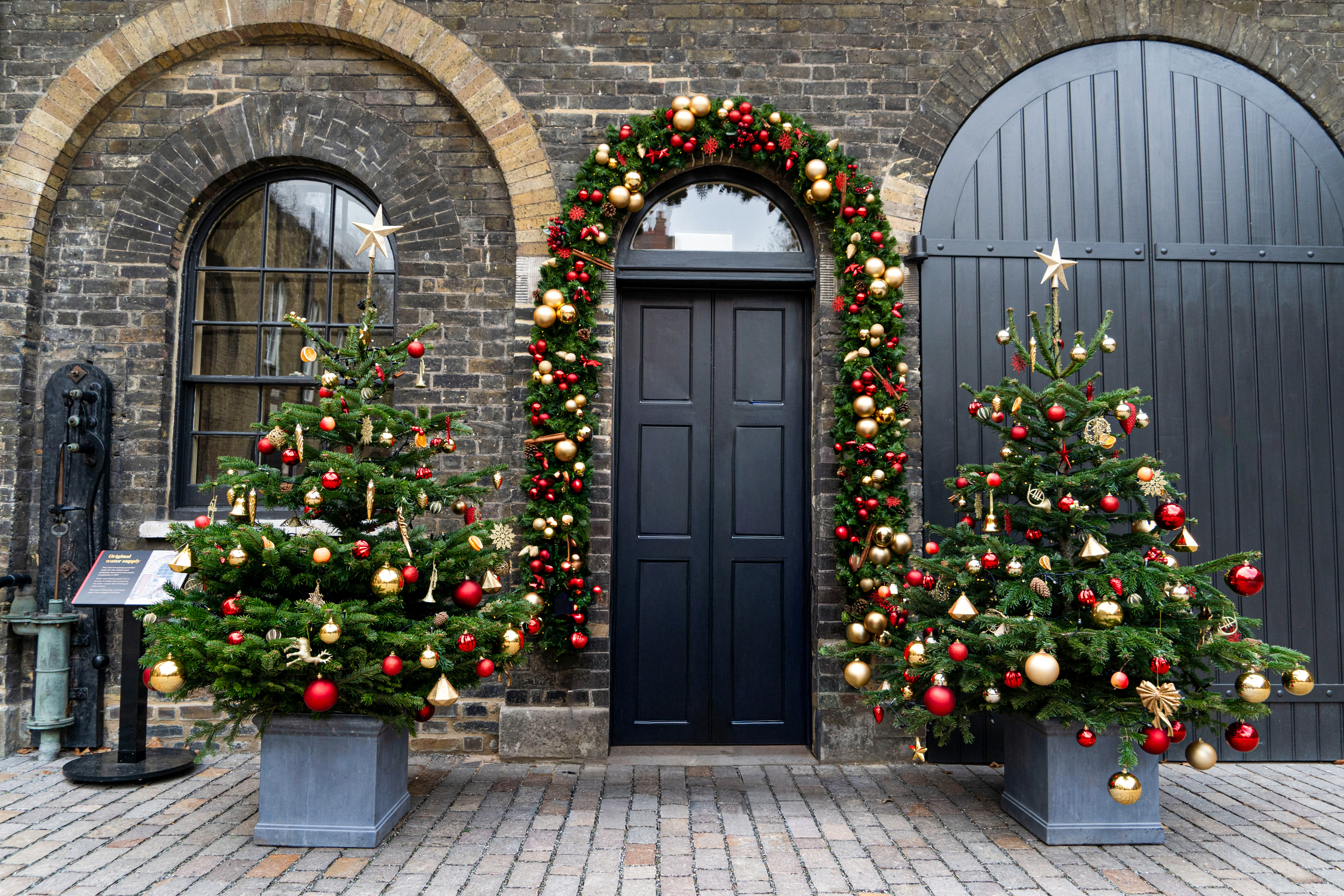 Two beautifully decorated gold, silver and red Christmas Trees stand in an outdoor courtyard besides a big black archway door with a festival garland surrounding. 