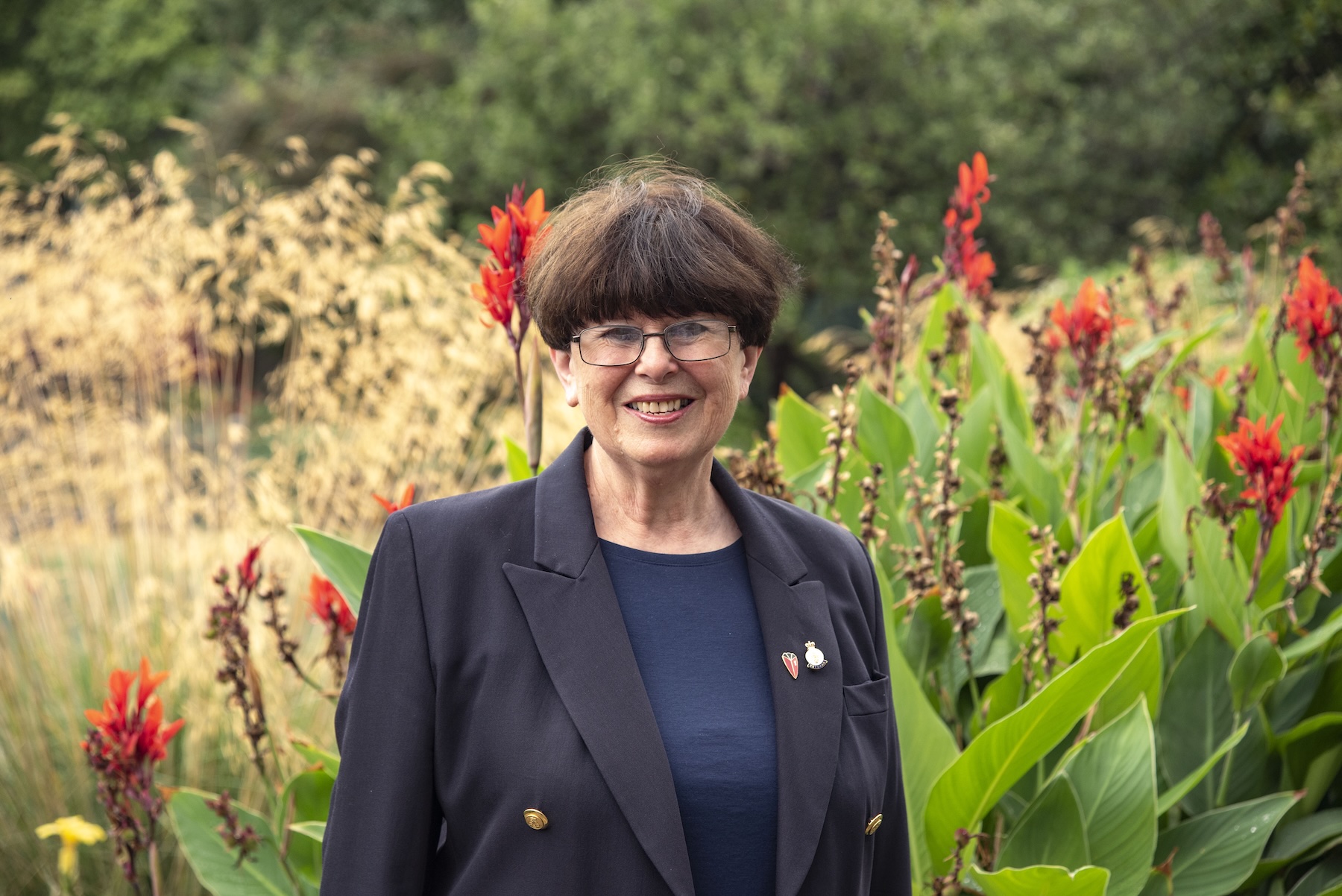 A lady with short dark hair and wearing glasses, stands in a wild garden with heavy foliage and red flowers. In contrast she's wearing a dark navy blazer and dark blue top with a Chelsea Pensioner lapel badge pinned to her jacket.