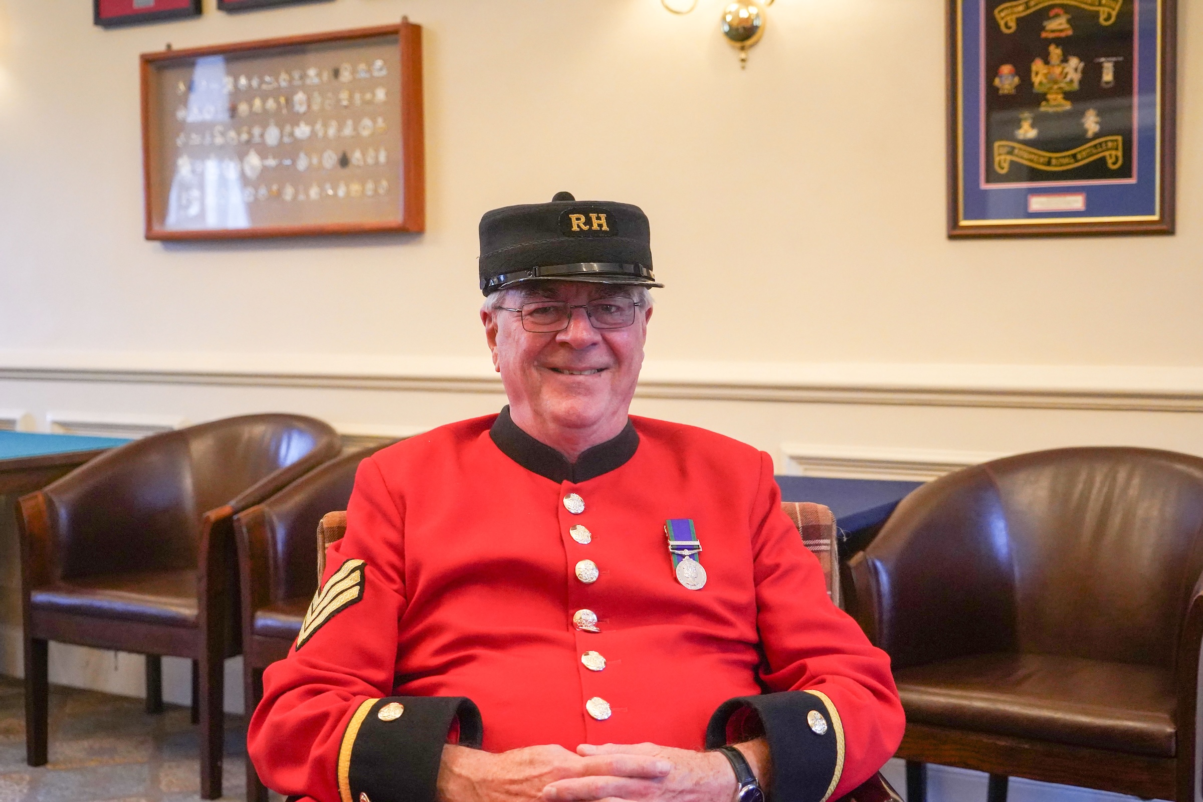 A male Chelsea Pensioner sits on a brown armchair, dressed in iconic scarlet uniform and black shako hat. A single medal adorn's his left breast.