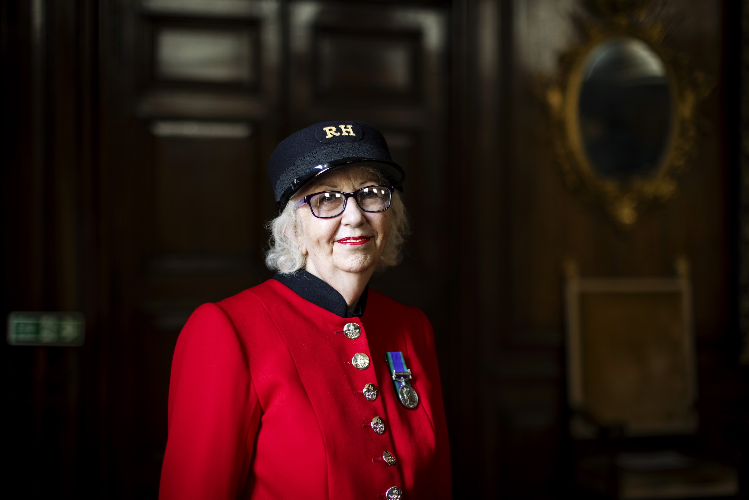 A female Chelsea Pensioner stands wearing her vibrant scarlet uniform, black shako hat and glasses, in front of large wood panelled doors.
