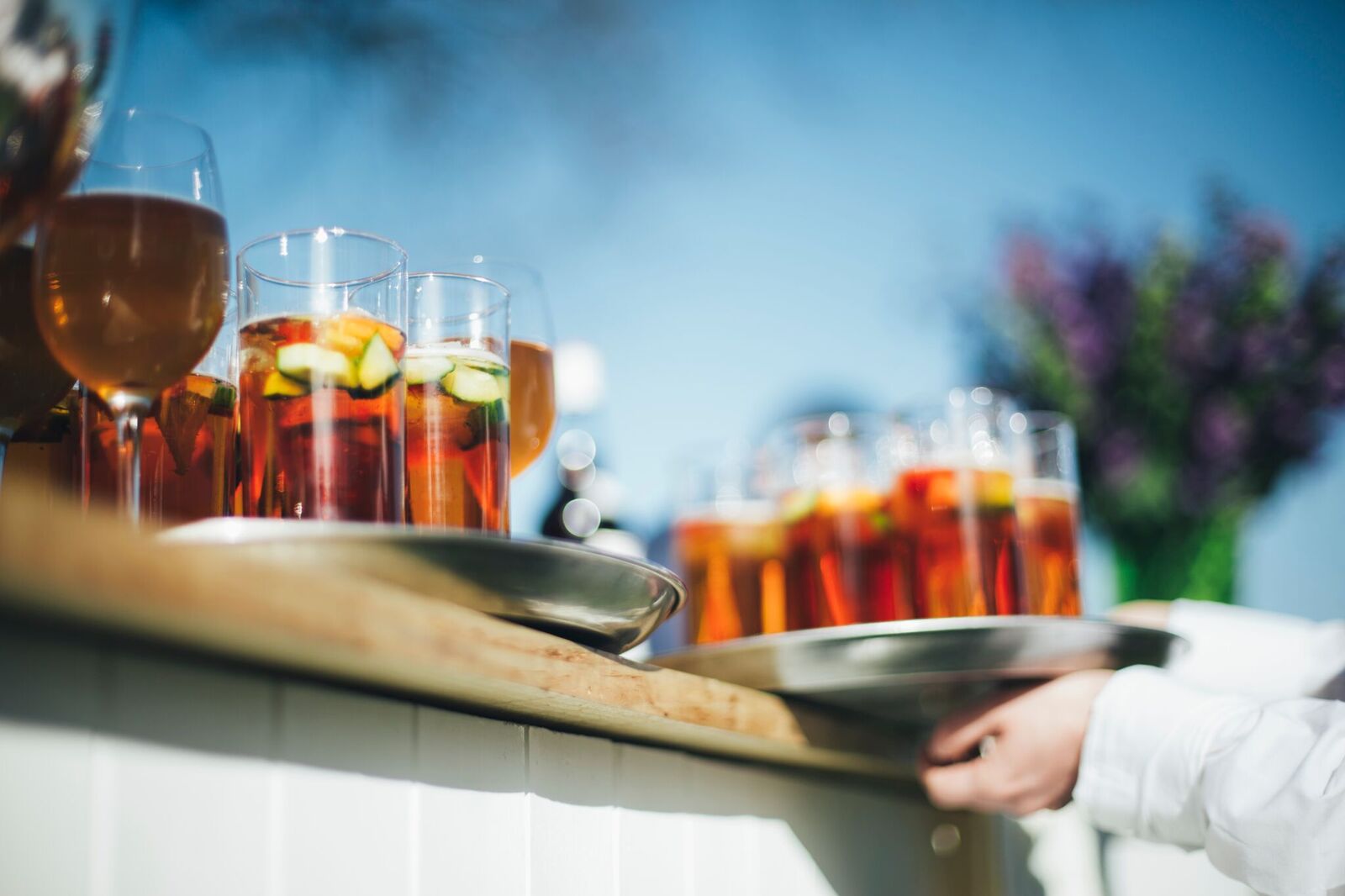 A close up view of trays filled with tall glasses of Pimm’s garnished with fruit and cucumber, set on an outdoor bar counter in bright sunshine. A server’s hand holds one of the trays. The background is softly blurred, showing a blue sky and a vase of flowers.