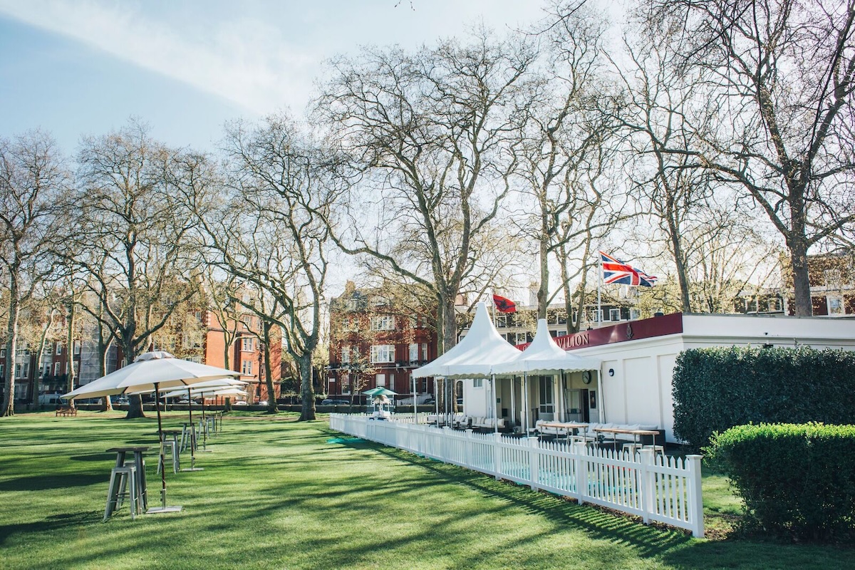 A bright outdoor scene showing the Pavilion at Burton Court, with white picket fencing, peaked canopy roofs, and flags flying above. The surrounding lawn is spacious and neatly kept, with rows of high tables and umbrellas set out on the grass. Tall leafless trees frame the background, along with red brick residential buildings under a clear sky.