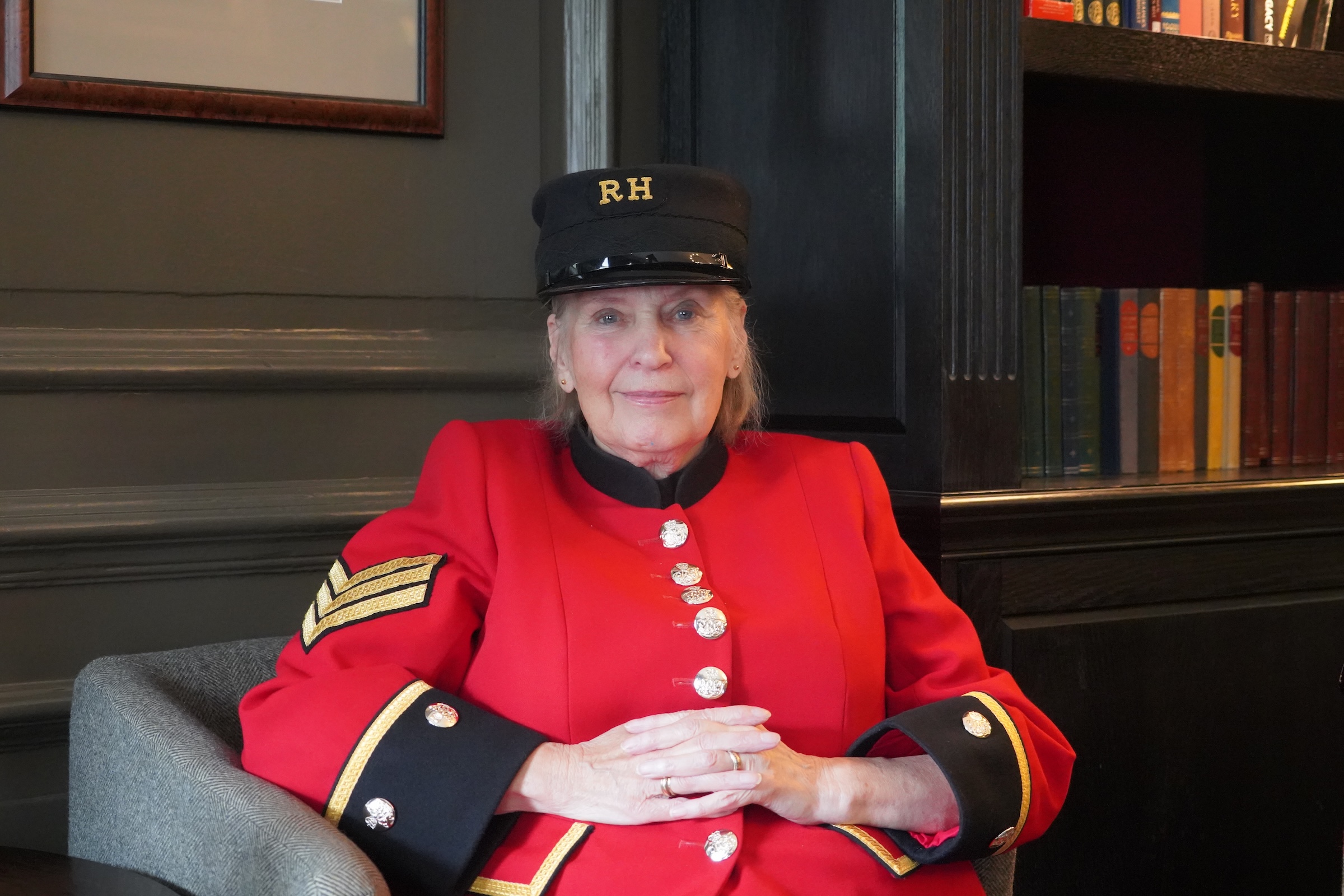 A lady Chelsea Pensioners sits within a wood panelled room in traditional scarlet uniform and black shako hat.