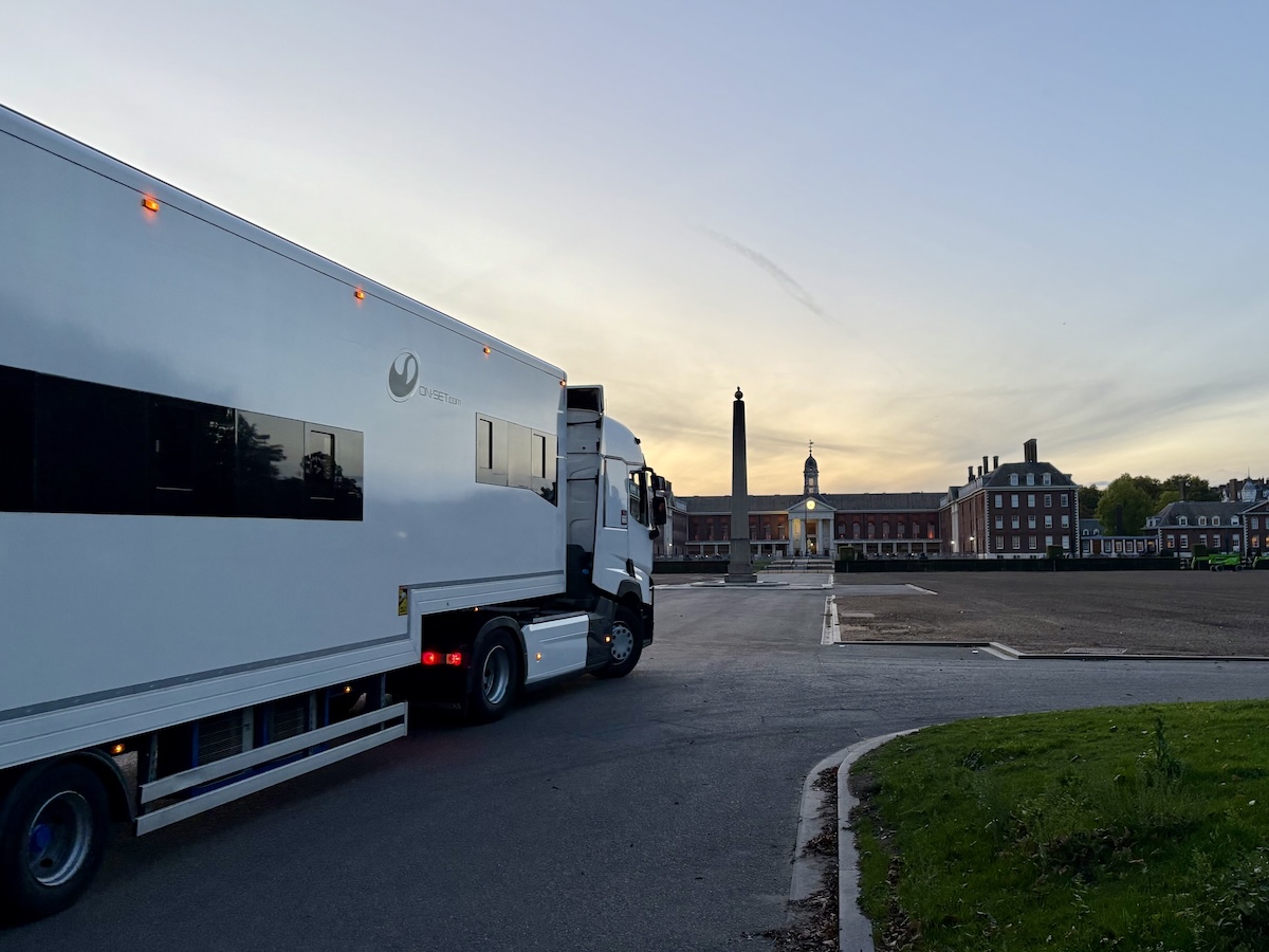 A large white production truck is parked at dawn near the grounds of the Royal Hospital Chelsea. The sky is pale with early morning light. In the distance, the Main Building and obelisk monument are visible. The scene is quiet, with empty paved areas and trimmed grass framing the approach to the historic buildings.