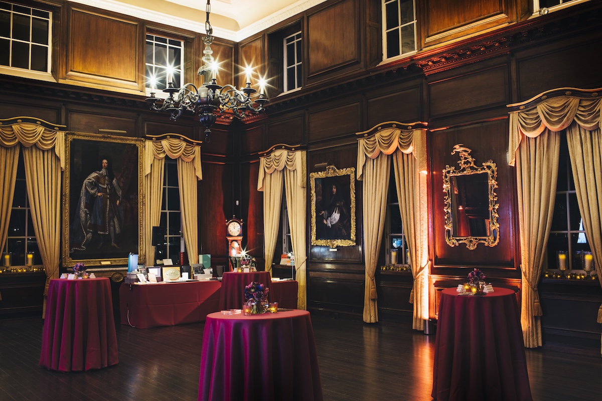 An opulent wood panelled room, with long flowing white curtains and red covered tables throughout the room. Large historic paintings hang on the walls and a warm light emphasises the night time shot.