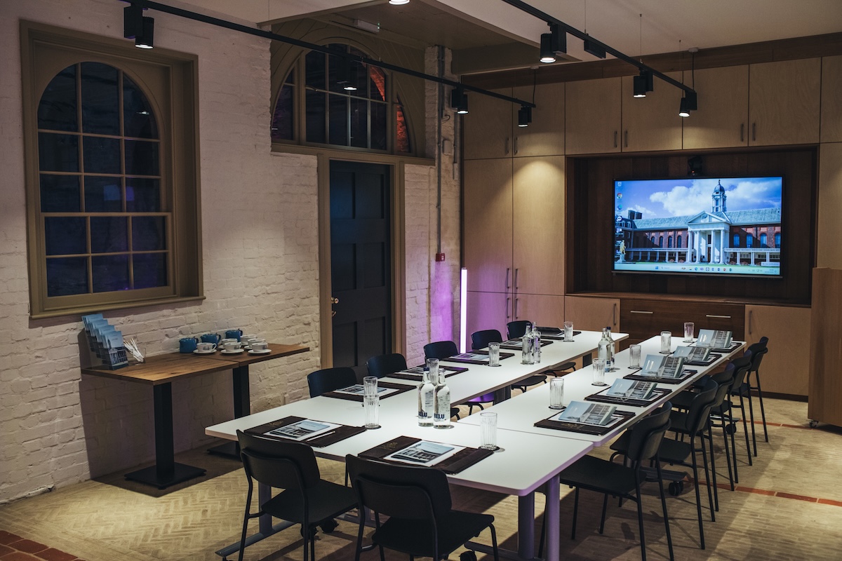 A modern meeting room with U shaped white tables set for a presentation. Each place has a glass, a notepad, and a bottle of water. Black chairs surround the tables. A large screen at the front displays an image of the Royal Hospital Chelsea. The room has exposed white brick walls, arched windows, warm overhead lighting, and built in wooden cabinetry. A side table holds stacked brochures and blue cups.