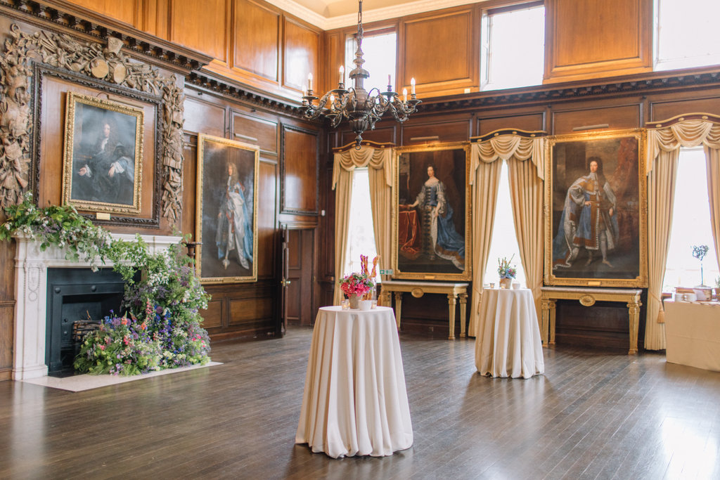 An elegant historic room with polished wooden floors, dark wood panelled walls, and large floor to ceiling portraits in ornate frames. Tall windows are dressed with gold curtains. A grand fireplace decorated with fresh floral arrangements sits on the left. Three round cocktail tables covered in white cloth stand in the centre of the room with small floral displays. A chandelier hangs overhead, adding to the refined and formal atmosphere.