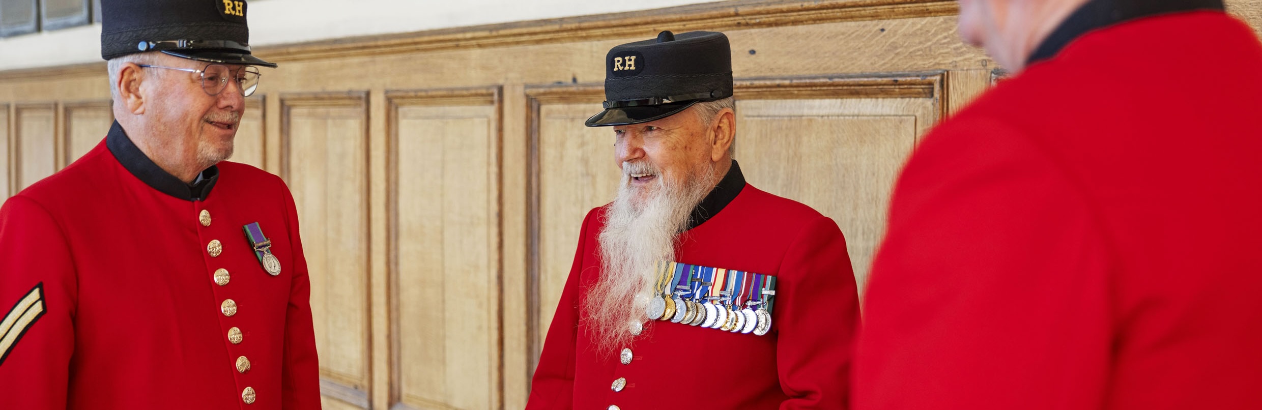 A group of Chelsea Pensioners stand in front of a wooden panelled wall, wearing their iconic scarlet uniforms with black Shako Hat emblazoned with gold "RH" lettering. The Pensioner in the centre has a long white beard and is highly decorated with almost a dozen medals.