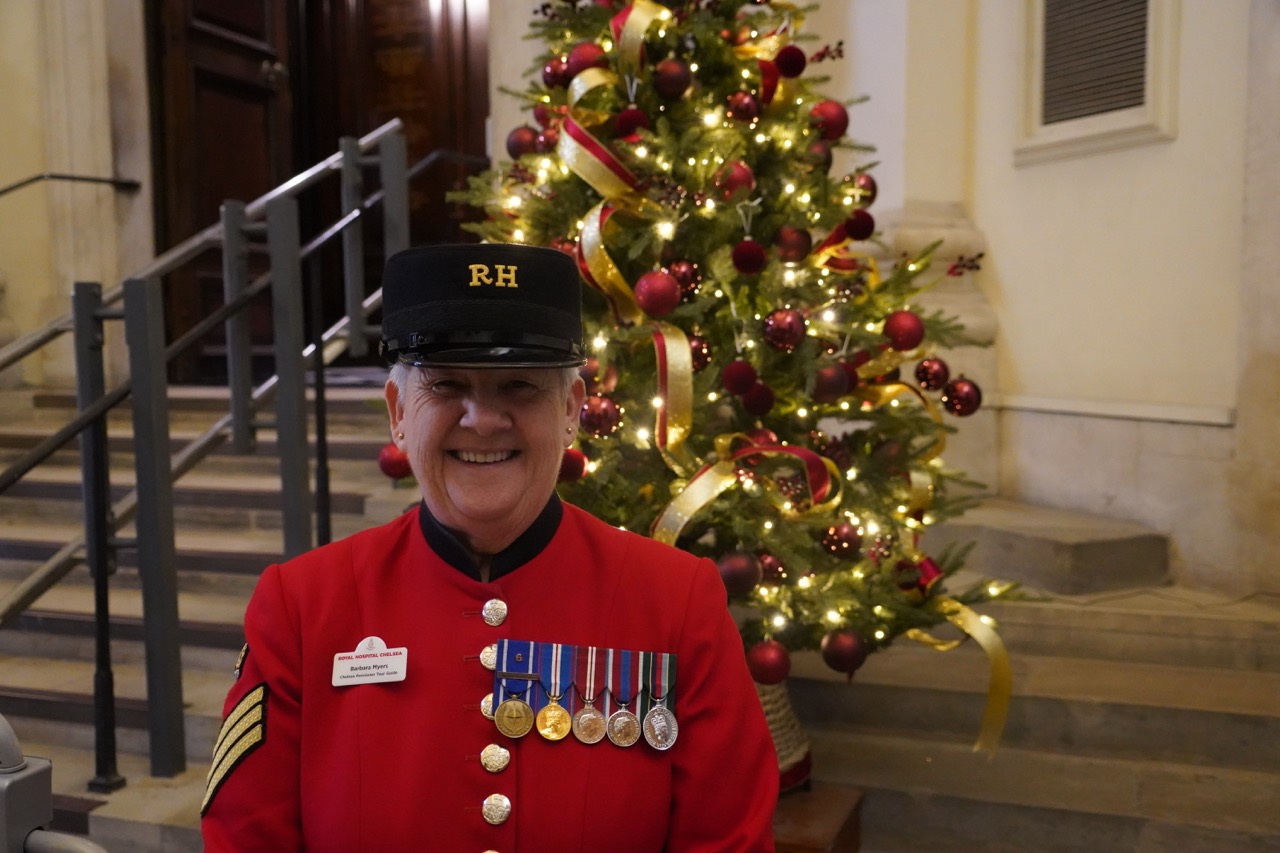A female Chelsea Pensioners stands in front of stone steps in the Royal Hospital Chelsea's Octagon foyer. Dressed in her famous Scarlet uniform and Shako hat, her name tag reads "Barbara - Tour Guide" her chest is adorned with half a dozen medals of various colours. Behind her a warmly decorated Christmas tree glitters in Red & Gold.