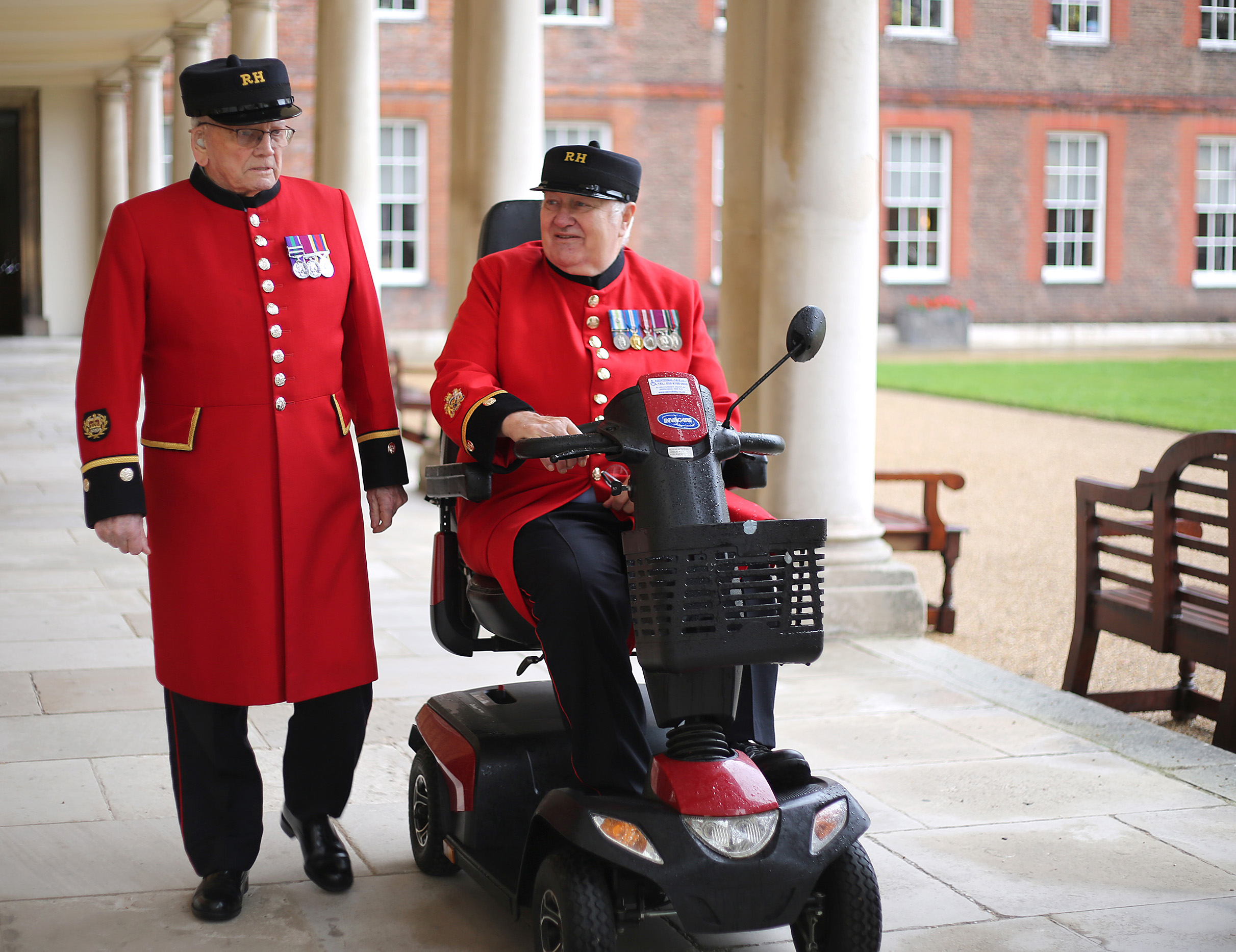 Two Chelsea Pensioners in scarlet uniforms walk together under the colonnade at the Royal Hospital Chelsea. One walks on foot while the other rides a mobility scooter, and they appear to be chatting. Both wear their RH caps and display service medals on their coats. The background shows the historic brick buildings and landscaped grounds.