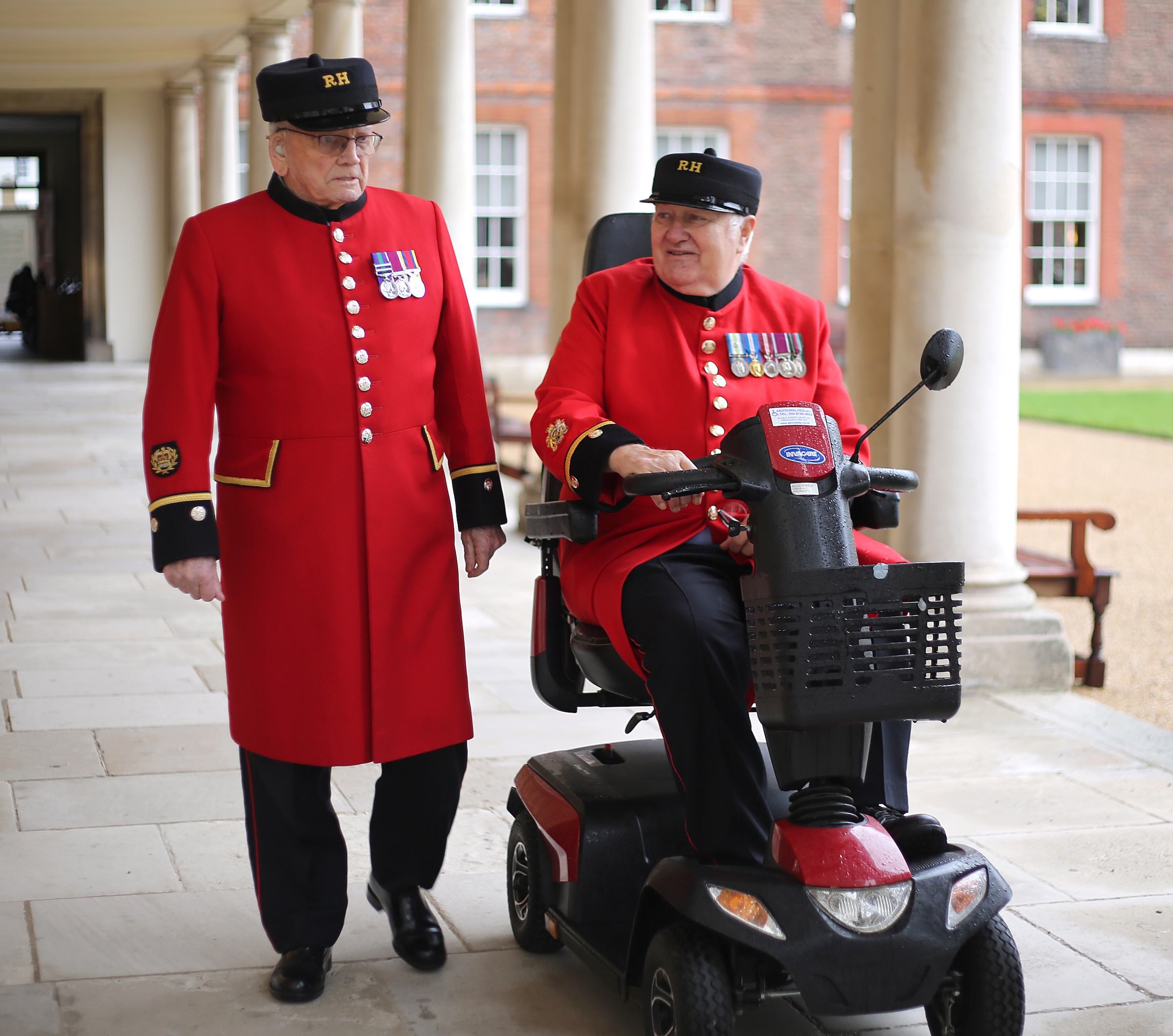 Two Chelsea Pensioners in scarlet uniforms walk together under the colonnade at the Royal Hospital Chelsea. One walks on foot while the other rides a mobility scooter, and they appear to be chatting. Both wear their RH caps and display service medals on their coats. The background shows the historic brick buildings and landscaped grounds.