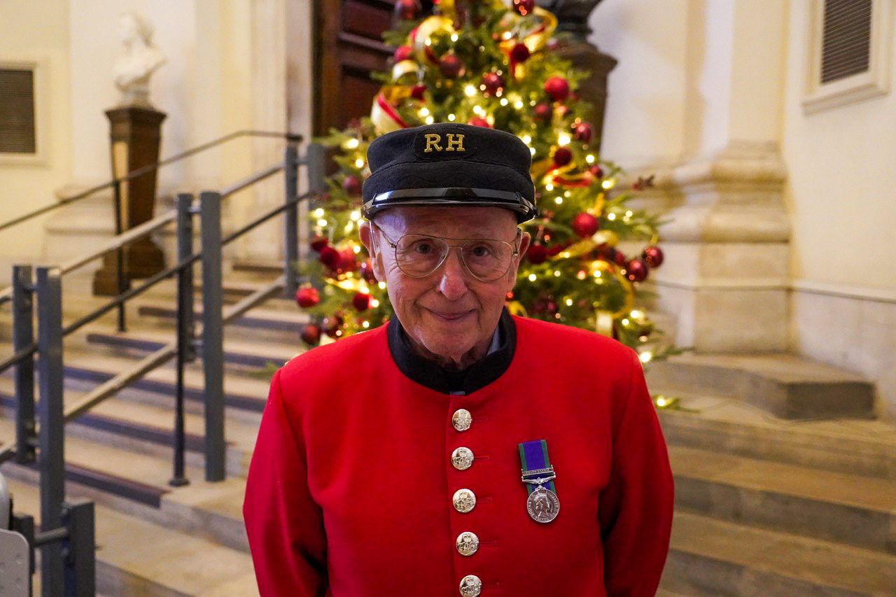 A Chelsea Pensioner in the traditional scarlet uniform and black cap stands indoors in front of a decorated Christmas tree. He wears glasses and has a single medal pinned to his chest. The background shows stone steps and warm festive lighting.