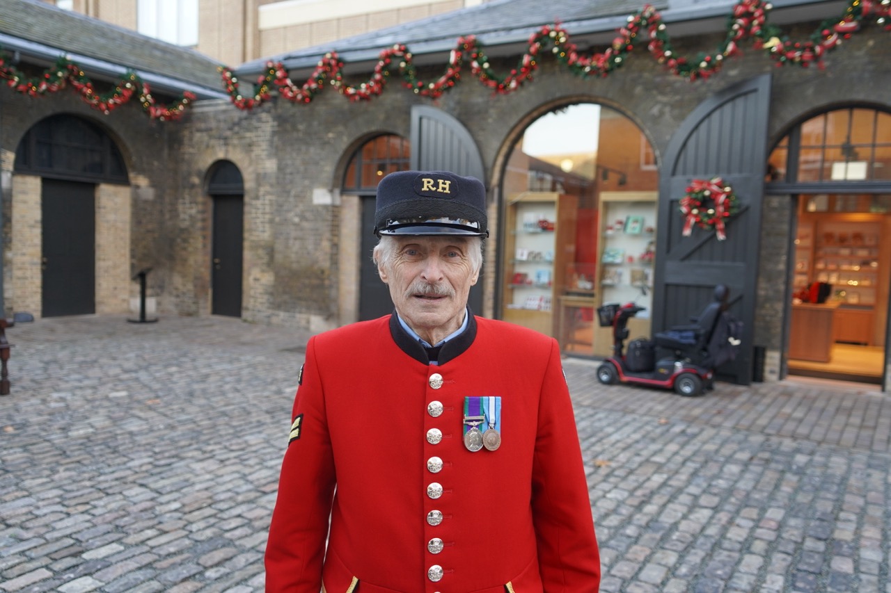 An elderly Chelsea Pensioner wearing the traditional bright scarlet uniform stands indoors in warm lighting. He has glasses, a white moustache, and a row of military medals pinned to his chest. Behind him are bookshelves with lamps and decorative flowers.