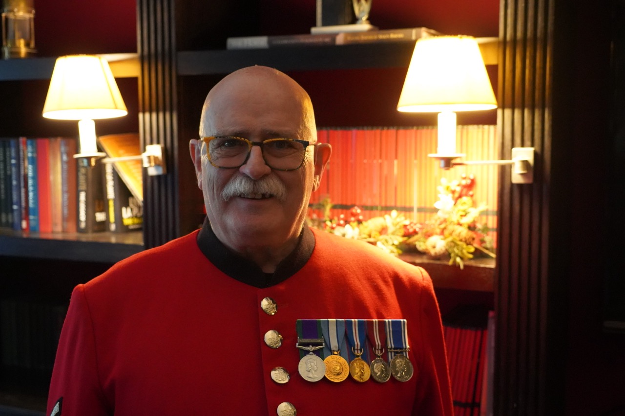 An elderly Chelsea Pensioner wearing the traditional bright scarlet uniform stands indoors in warm lighting. He has glasses, a white moustache, and a row of military medals pinned to his chest. Behind him are bookshelves with lamps and decorative flowers.