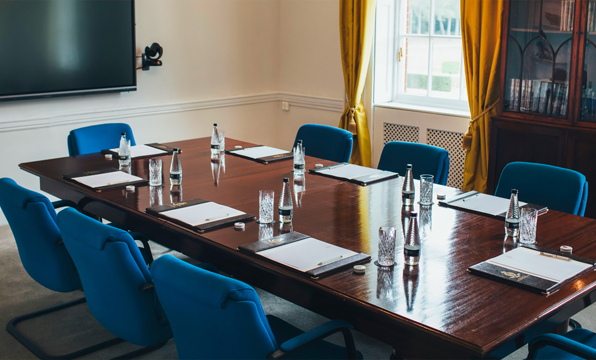 A polished wooden boardroom table surrounded by modern blue chairs, each place set with a notepad, pen, water bottle, and glass. A large flat-screen monitor is mounted on the wall, and sunlight streams through a window with gold curtains. A traditional wooden bookcase filled with books sits in the corner, blending modern meeting facilities with classic elegance.