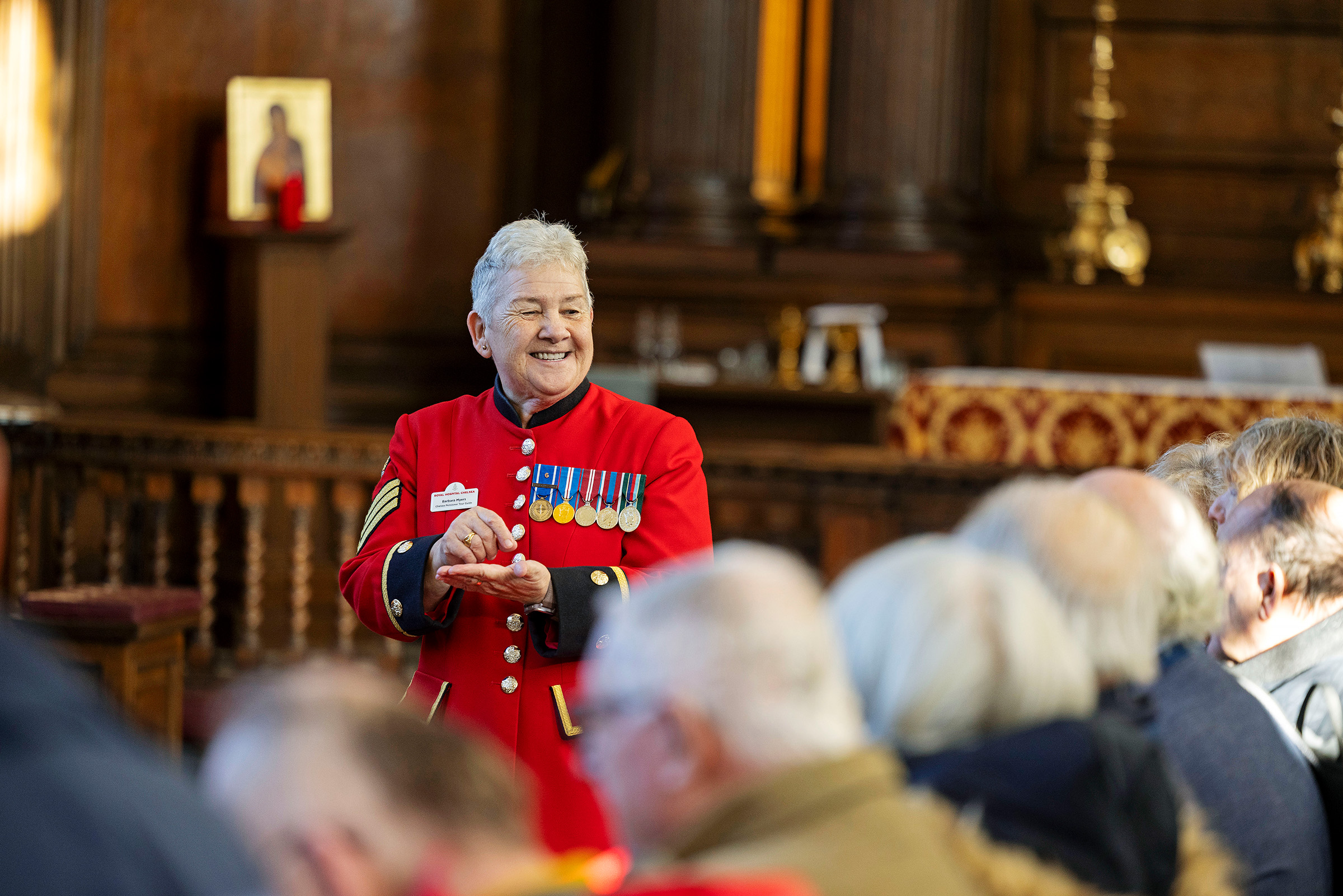 Chelsea Pensioner in a scarlet uniform with medals addressing a seated audience inside the chapel at the Royal Hospital Chelsea.