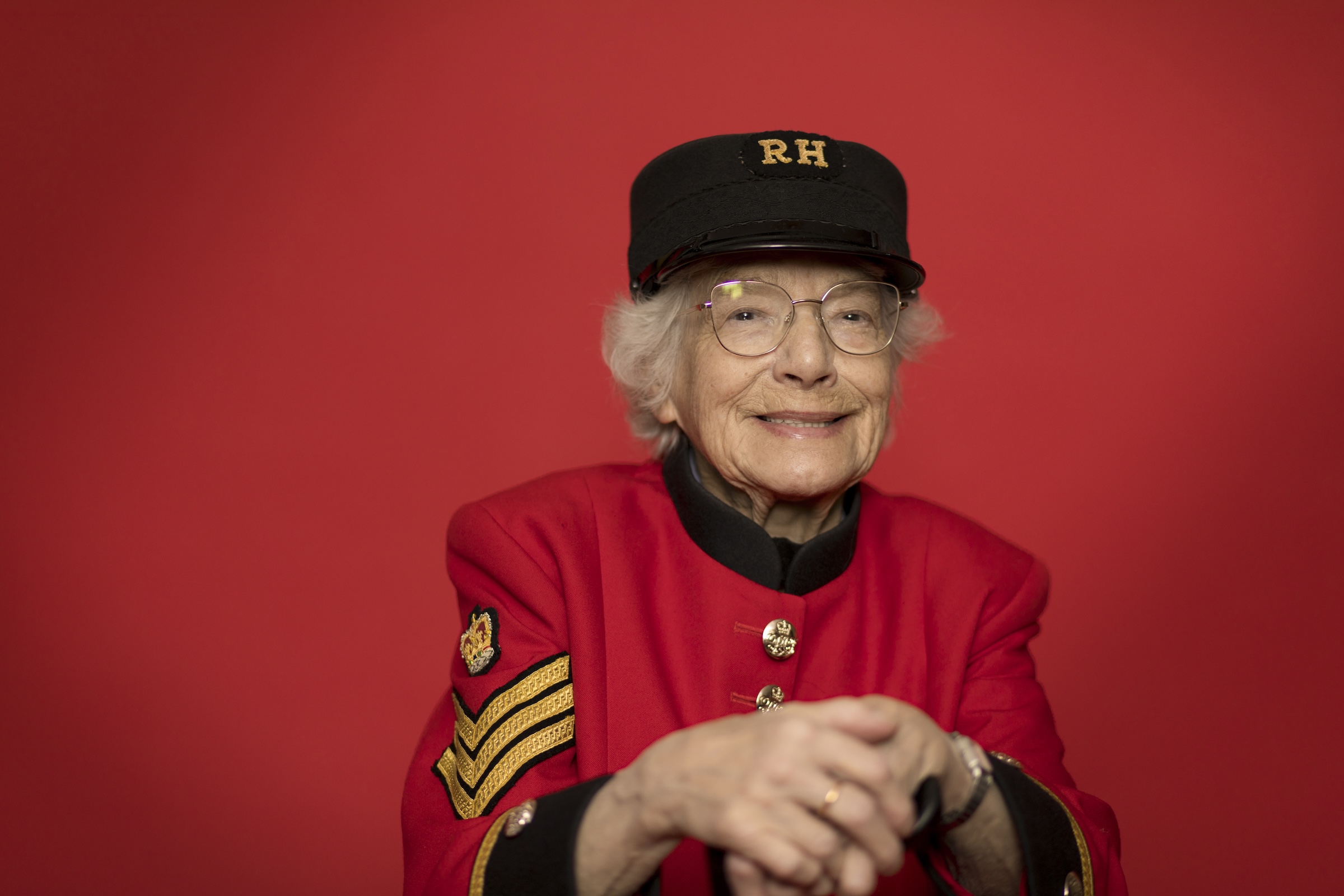 Elderly Chelsea Pensioner seated against a red backdrop, wearing a scarlet uniform with service chevrons and an RH cap, hands folded.
