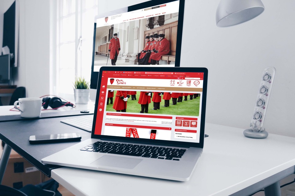 A laptop screen and desktop monitor are open and displaying various Chelsea Pensioner imagery, set upon a grey clean minimalist desk