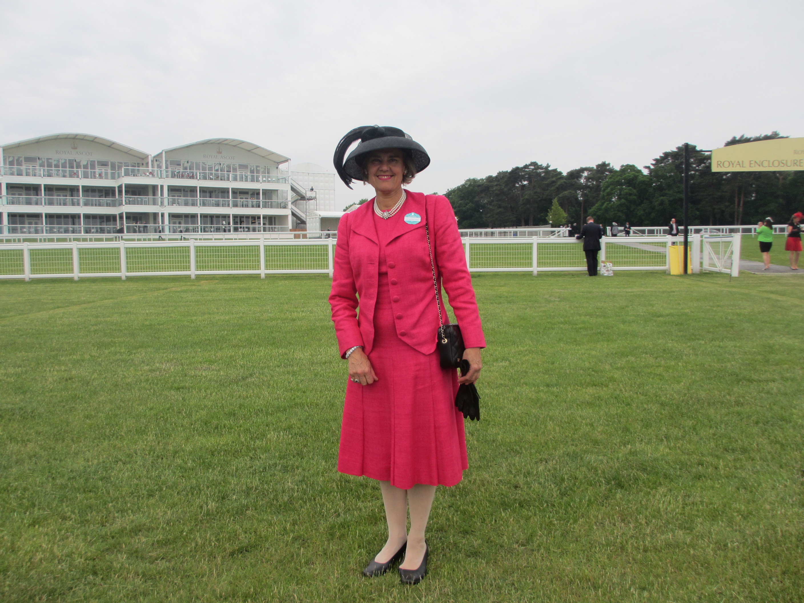 A lady stands on the grounds of a horse racing track, dressed in a bright pink dress suit and black hat and holding a black pair of gloves and handbag.