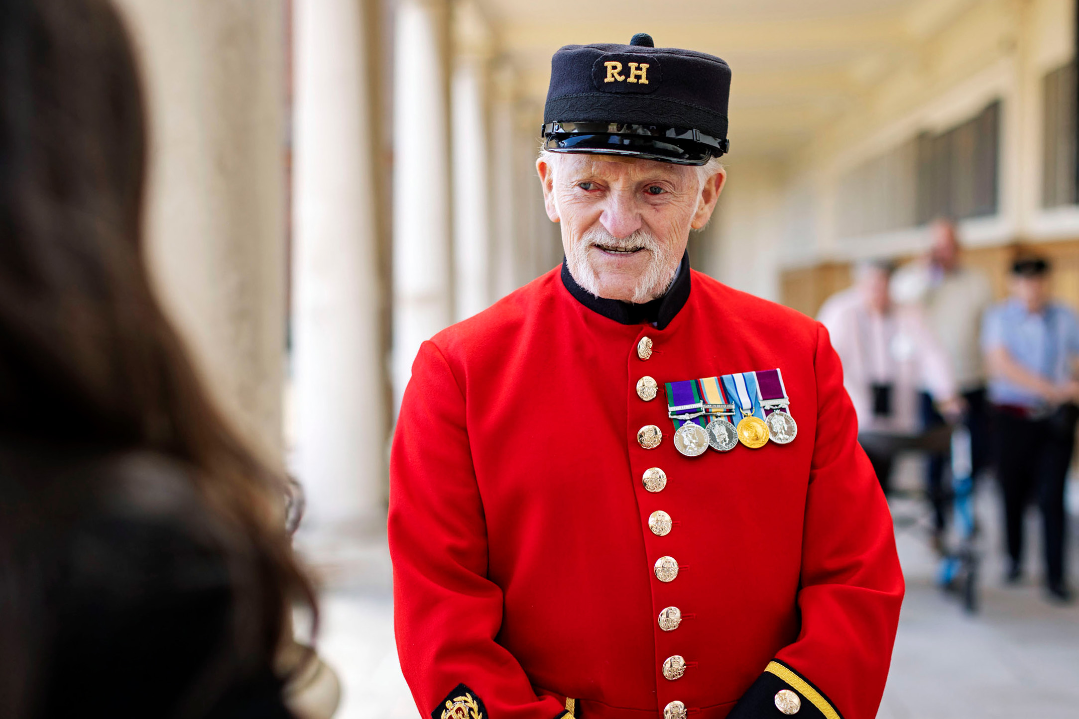 A Chelsea Pensioner in scarlet uniform with medals speaks to a visitor in a colonnaded walkway at the Royal Hospital Chelsea.