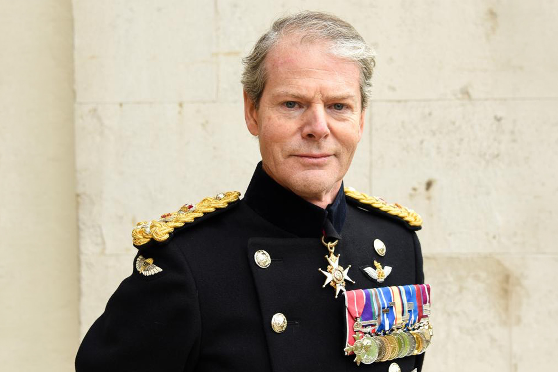 A highly decorated 4-star General of the British Army stands with hands behind his back against a white brick building