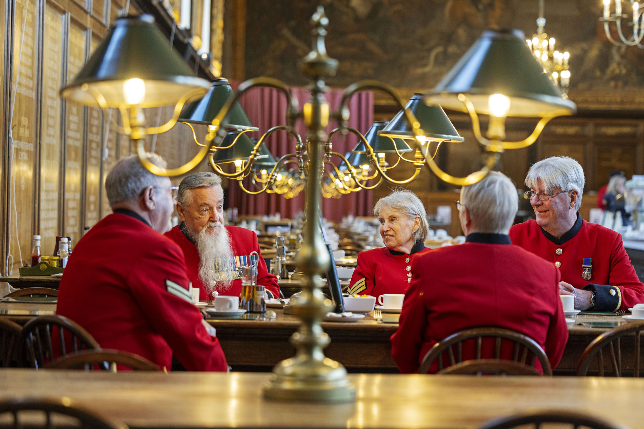 Chelsea Pensioners in scarlet uniforms sit together talking over tea in the historic dining hall at the Royal Hospital Chelsea.