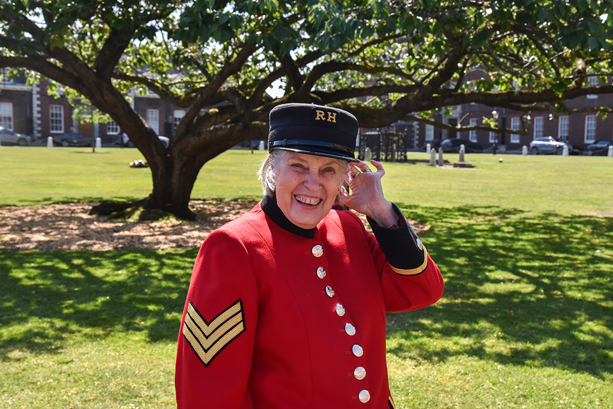 A smiling Chelsea Pensioner in scarlet uniform stands in the grounds of the Royal Hospital Chelsea beneath a tree.