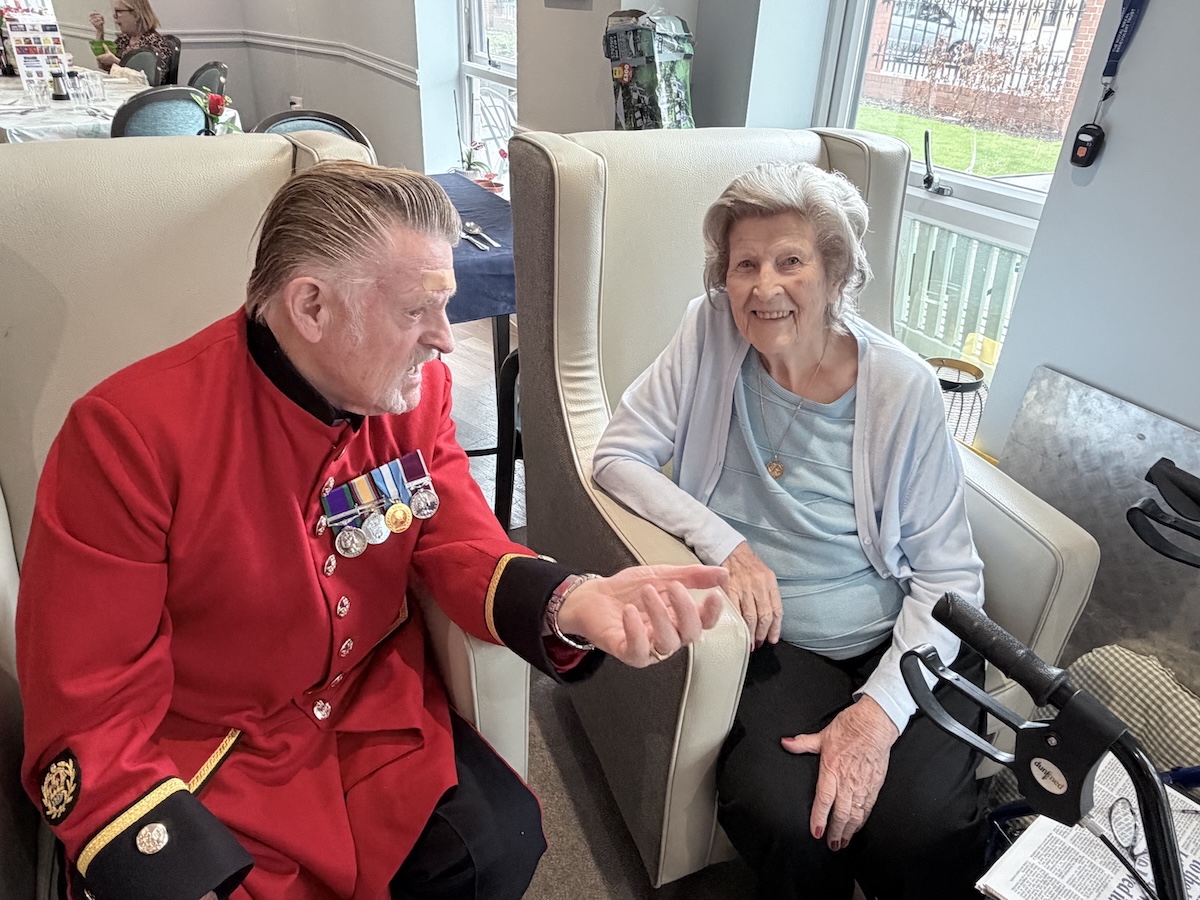 A Chelsea Pensioner sits dressed in his iconic scarlet uniform alongside a female veteran in a warmly lit room