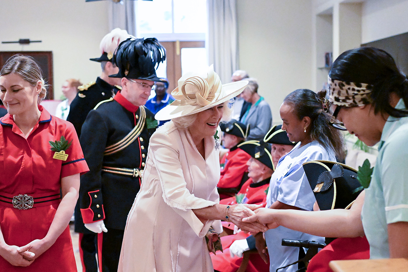 Queen Camilla, dressed in a pale cream outfit and wide brimmed hat, greets healthcare staff and residents during a visit to the Royal Hospital Chelsea, while Chelsea Pensioners and ceremonial guards look on in the background.