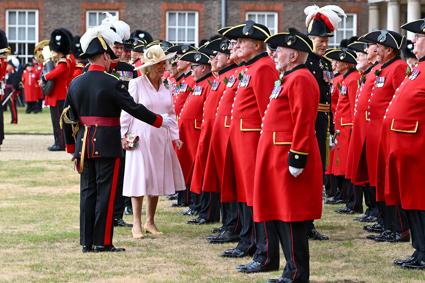 Queen Camilla walks along a line of Chelsea Pensioners in scarlet uniforms during a ceremonial parade at the Royal Hospital Chelsea, accompanied by military officers in full dress uniform as she speaks with participants.