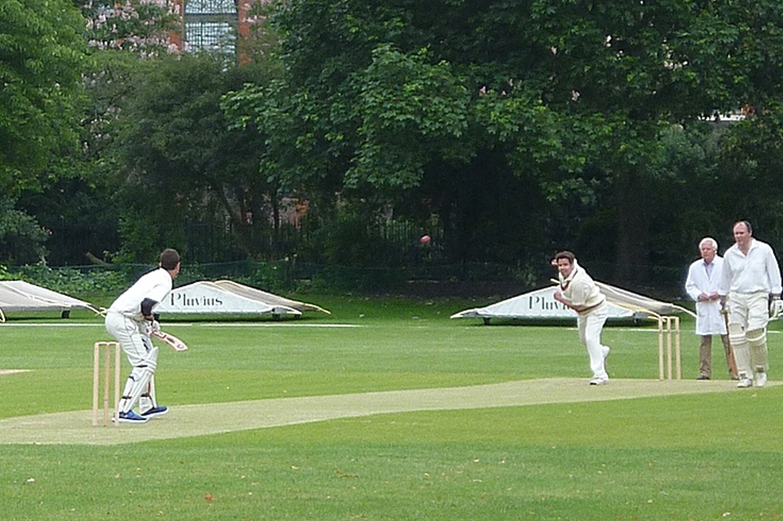 Players competing in a cricket match on the Royal Hospital Chelsea’s historic Burton Court cricket ground, surrounded by mature trees and the Hospital’s heritage buildings.