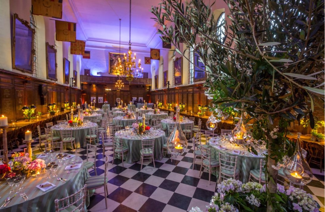 A photo of the Great Hall at the Royal Hospital Chelsea, dressed for an event with white clothed round tables, atmospheric lighting and a centrepiece indoor tree showcasing the space