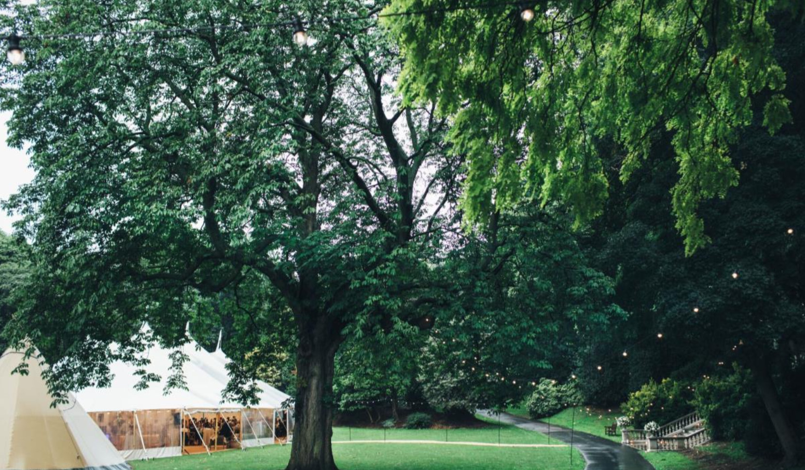 An elegant wedding marquee set within the Ranelagh Gardens at the Royal Hospital Chelsea, surrounded by mature trees, illuminated festoon lighting, landscaped lawns and the historic stone staircase.