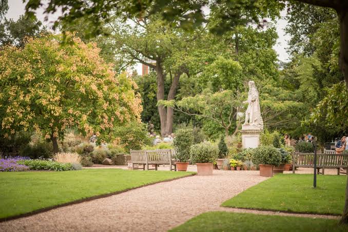 A stone statue on a pedestal stands amid potted shrubs and lawns, flanked by park benches along a gravel path, with mature trees in the background.