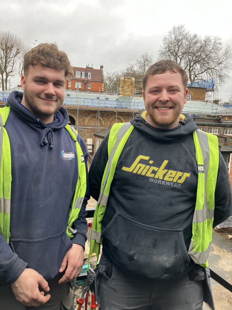 Two electrical apprentices in hi-vis jackets stood smiling in front of building site.