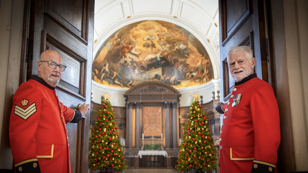 Two Chelsea Pensioners, dressed in their iconic scarlet uniforms, stand either side of large wooden doors, which are partially opened revealing a historic chapel, with Fresco painted mural, wooden altar and two Christmas trees either side.