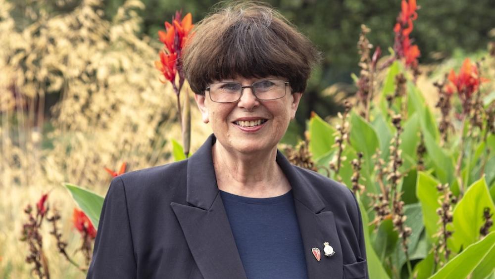 A lady with short dark hair and wearing glasses, stands in a wild garden with heavy foliage and red flowers. In contrast she's wearing a dark navy blazer and dark blue top with a Chelsea Pensioner lapel badge pinned to her jacket.