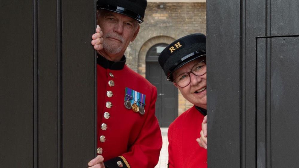 Two Chelsea Pensioners, dressed in their iconic Scarlet Coats and black shako hats peek out from behind partially opened large black doors