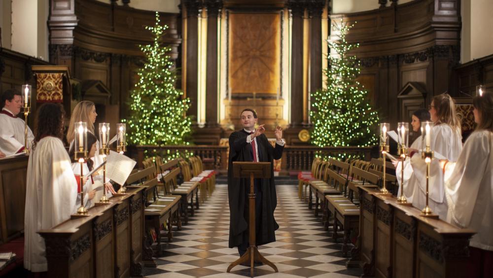 A choir performs inside a historic chapel decorated for Christmas. A conductor stands at a wooden lectern in the centre of a black and white chequered aisle, directing singers dressed in white robes who stand on either side holding candles and sheet music. Two illuminated Christmas trees flank the ornate wooden altar at the back of the chapel, creating a warm and festive atmosphere.